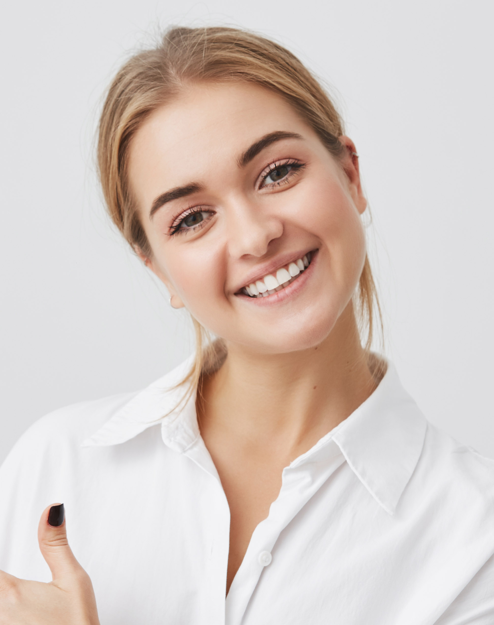 excited pretty young blonde caucasian woman wearing white shirt pointing her index finger sideways, smiling joyfully with her white teeth, showing something surprising on gray copy space wall